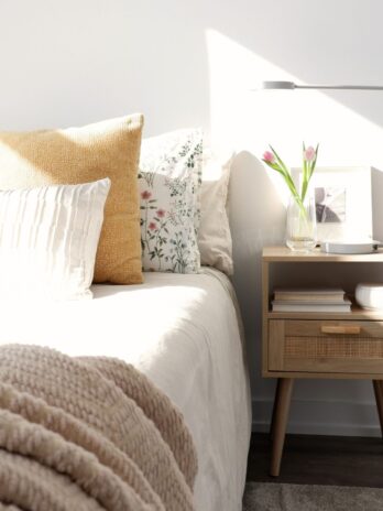A bright bedroom corner showing a bed with layers of neutral blankets, textured mustard yellow and floral pillows, next to a light wood nightstand with a vase of pink tulips.