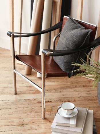 A modern chair with leather seat and metal frame sits on a wooden floor, topped with a textured gray cushion. Nearby is a stack of books with a teacup on top, and a plant and rolled canvases in the background.