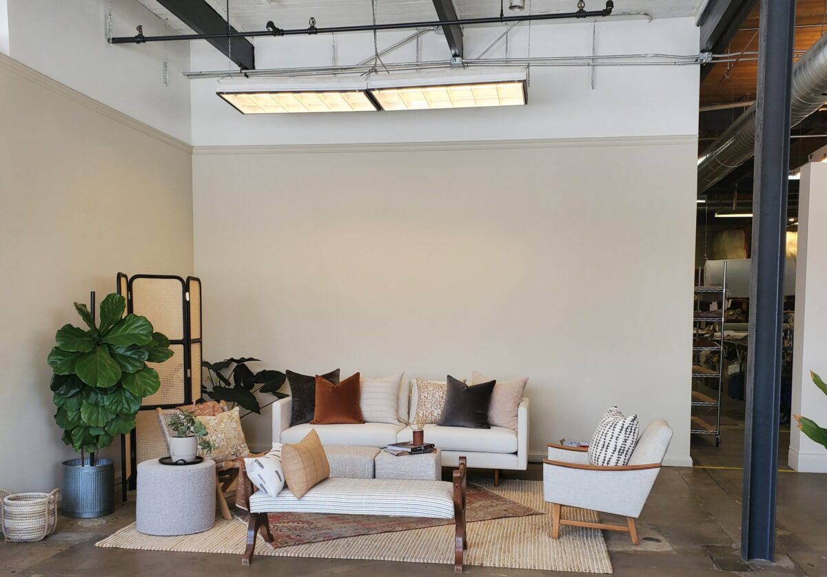 A modern living room with a white sofa, various throw pillows, two armchairs, a bench, potted plants, a wooden room divider, and neutral-colored decor on a woven rug under bright ceiling lights.