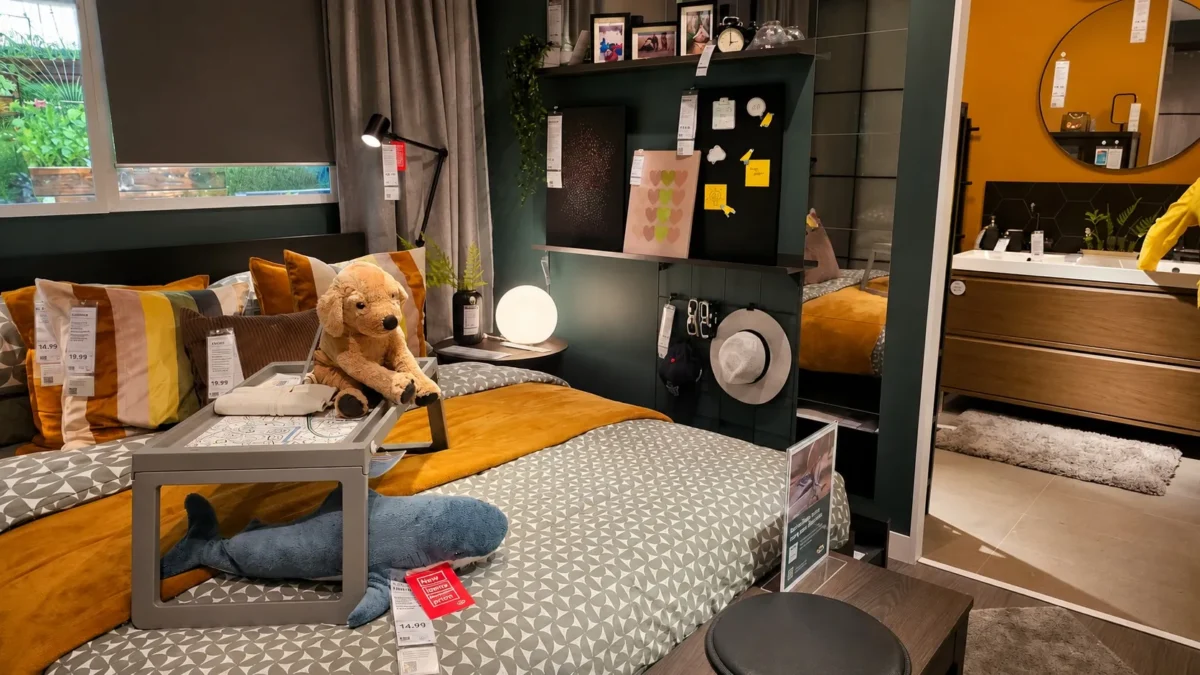 A cozy bedroom display features a bed with yellow and gray bedding, a tray, a stuffed dog and shark, shelves with photos above the bed, and a view into a modern bathroom with a large mirror.