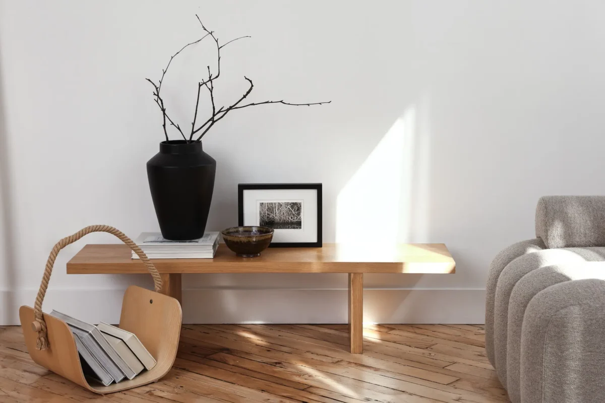 A minimalist living space with a wooden bench holding a black vase with bare branches, a framed photo, and a bowl. Nearby, a wooden magazine holder contains books and a beige sofa sits on a wooden floor.
