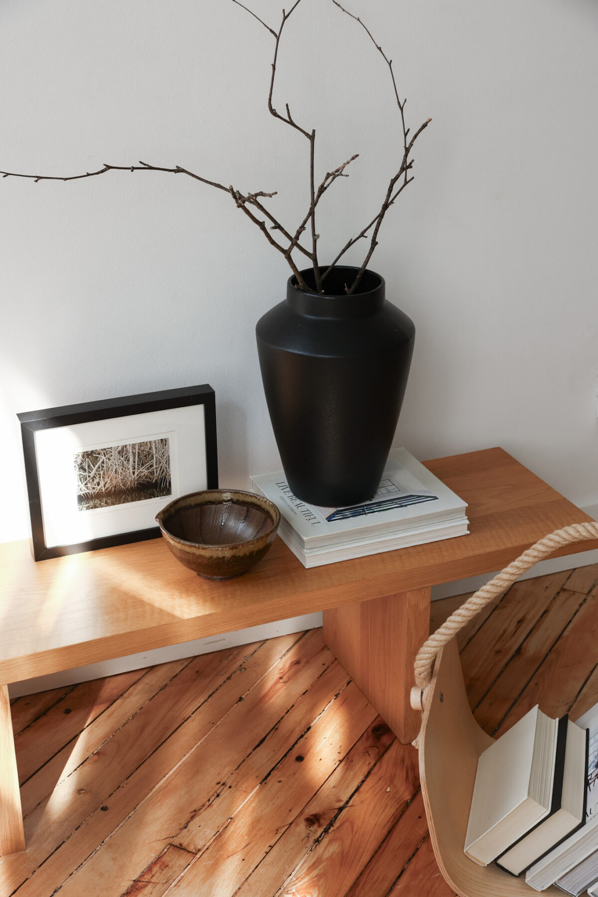 A minimalist wooden bench displays a black vase with bare branches, a small brown bowl, stacked books, and a framed nature photo. A wooden basket with a rope handle and books sits on a wood floor nearby.