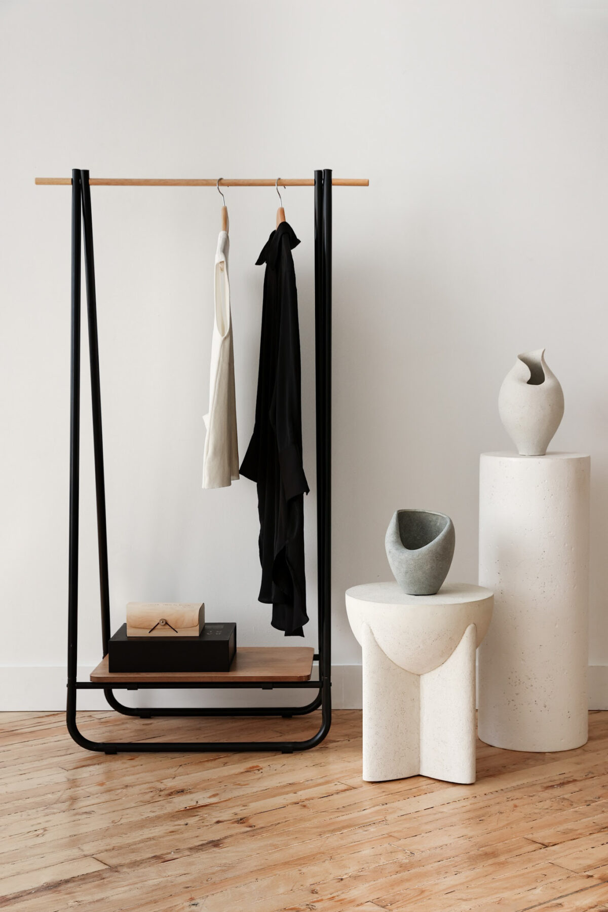 Minimalist interior with a black clothing rack holding a white top and a black garment, next to white sculptural vases on cylindrical and geometric pedestals, set on a wooden floor against a plain white wall.