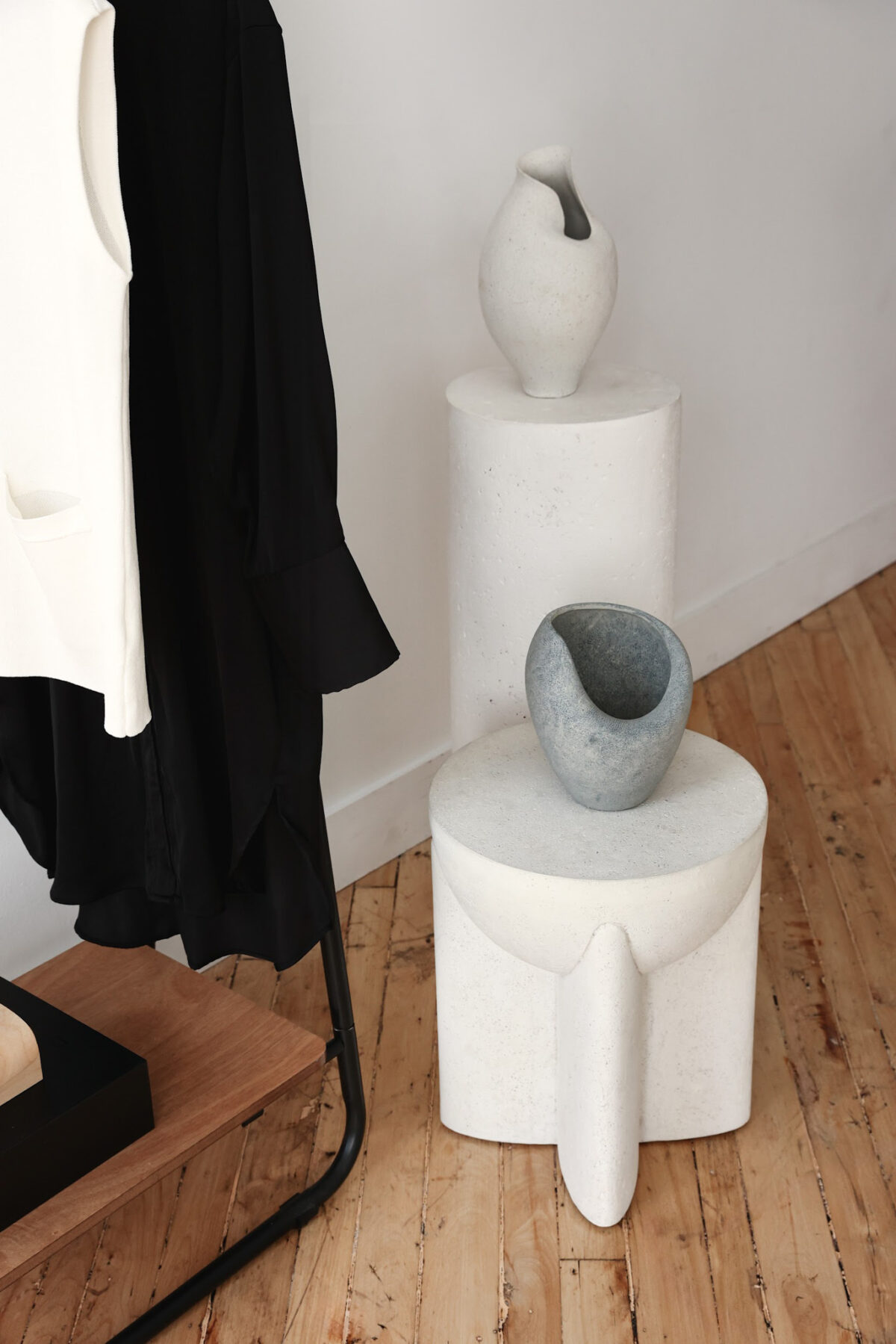 Two modern ceramic vases sit on white sculptural pedestals by a wooden floor. A black and a white garment hang on a rack nearby, creating a minimalist and stylish interior scene.