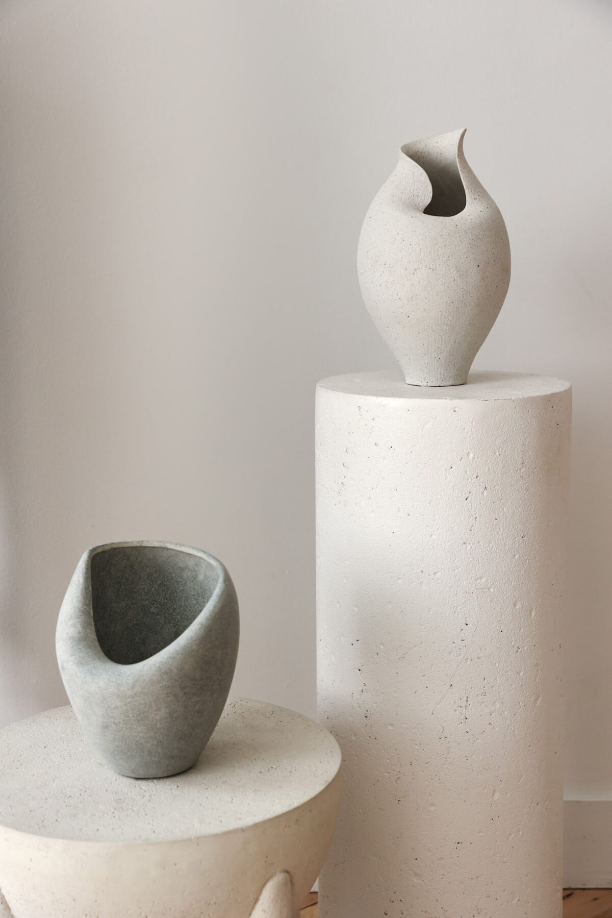 Two sculptural, minimalist ceramic vases with organic, asymmetrical shapes rest on textured, cylindrical white pedestals against a neutral wall. The vases are gray and off-white in color.