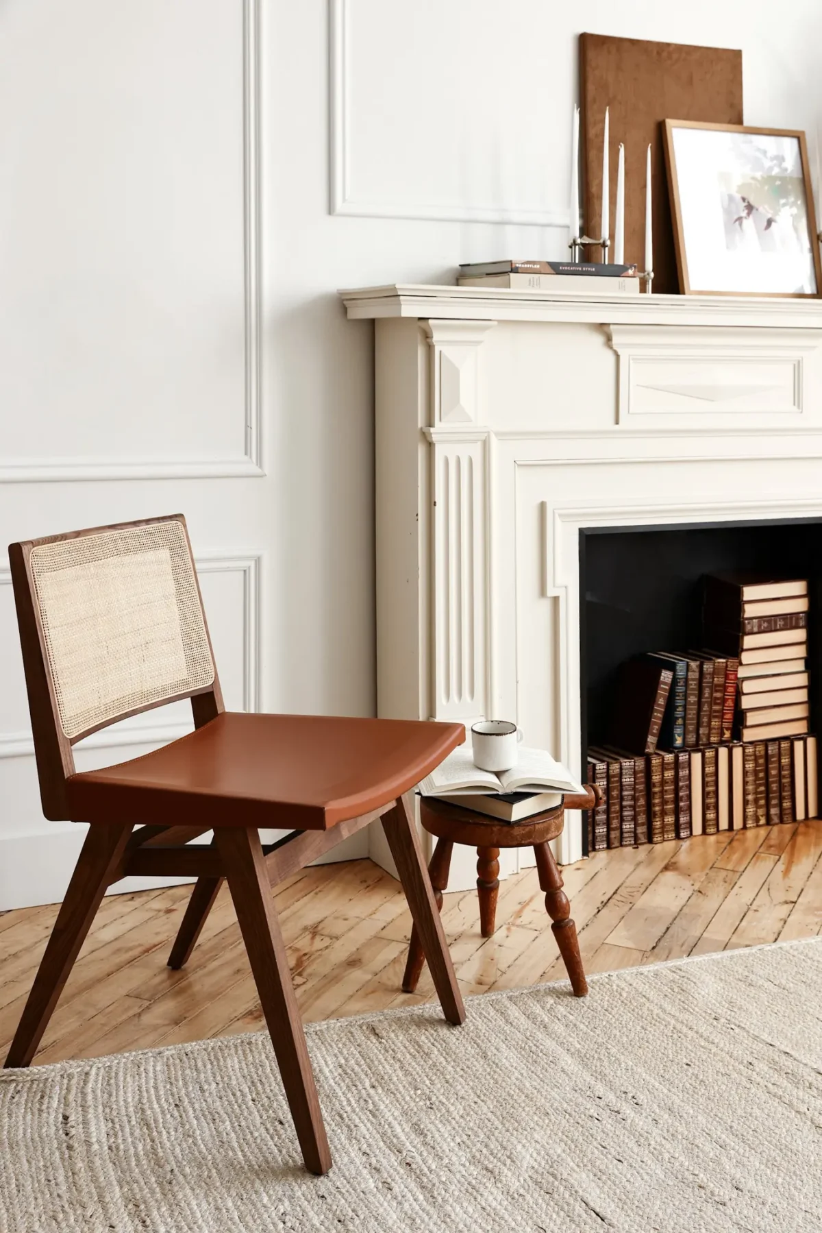 A wooden chair with a woven backrest sits beside a small round stool holding a cup and book, in front of a white ornate fireplace filled with stacked books. The room has wooden floors and neutral décor.