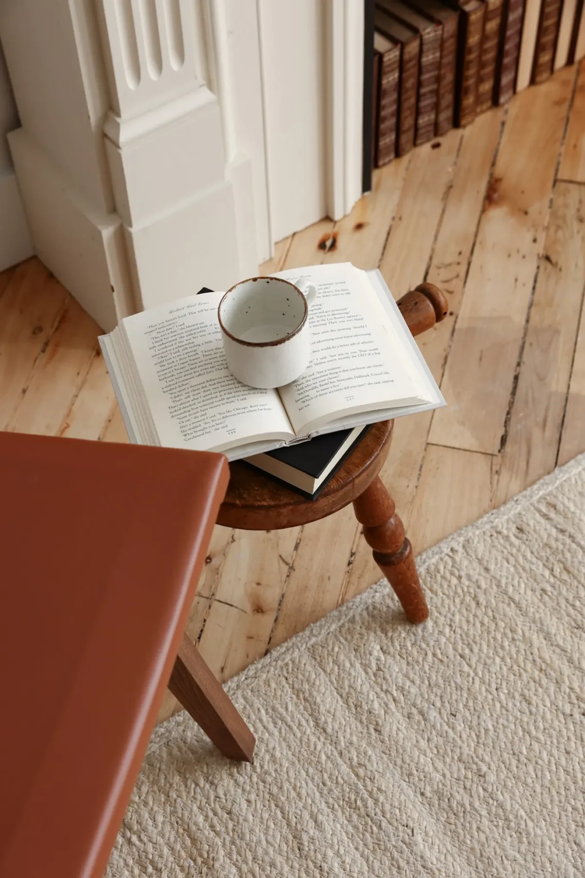 A ceramic mug sits on top of an open book placed on a small wooden stool next to a textured cream rug, with bookshelves and wooden floorboards in the background.