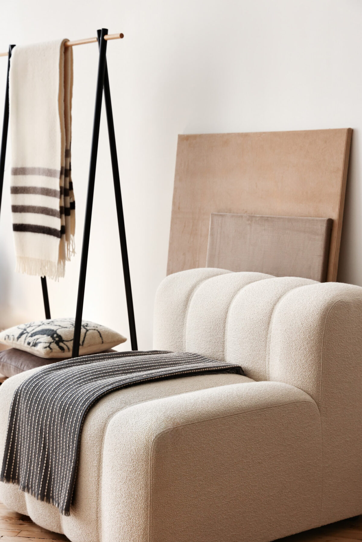 A modern, cream-colored, textured sofa with a dark striped throw blanket draped over it. Behind, there is a wooden bench, striped blanket on a black frame rack, and large wooden panels leaning against a white wall.