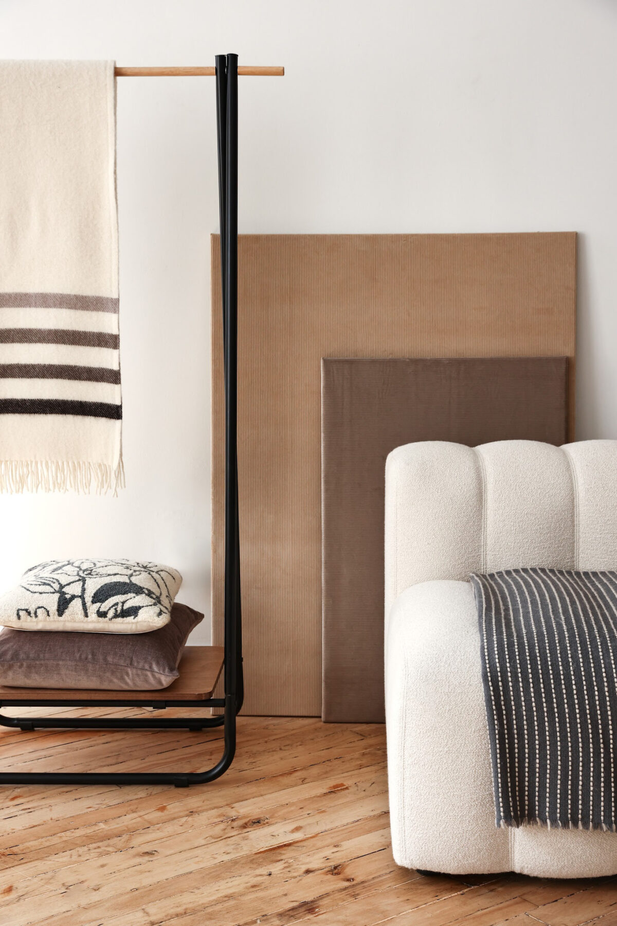 A cozy modern room with a white upholstered sofa, a striped blanket draped over the arm, a black metal rack with a striped throw, stacked pillows, and neutral-toned panels against a white wall on a wooden floor.