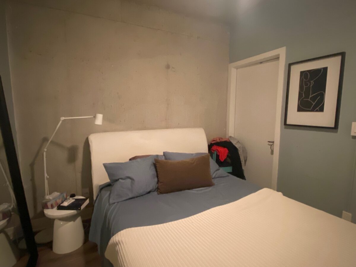 A cozy bedroom with a bed against a concrete wall, blue and brown bedding, a white side table with a lamp, a clothes pile on the chair, and abstract art on a pale blue wall near a closed door.