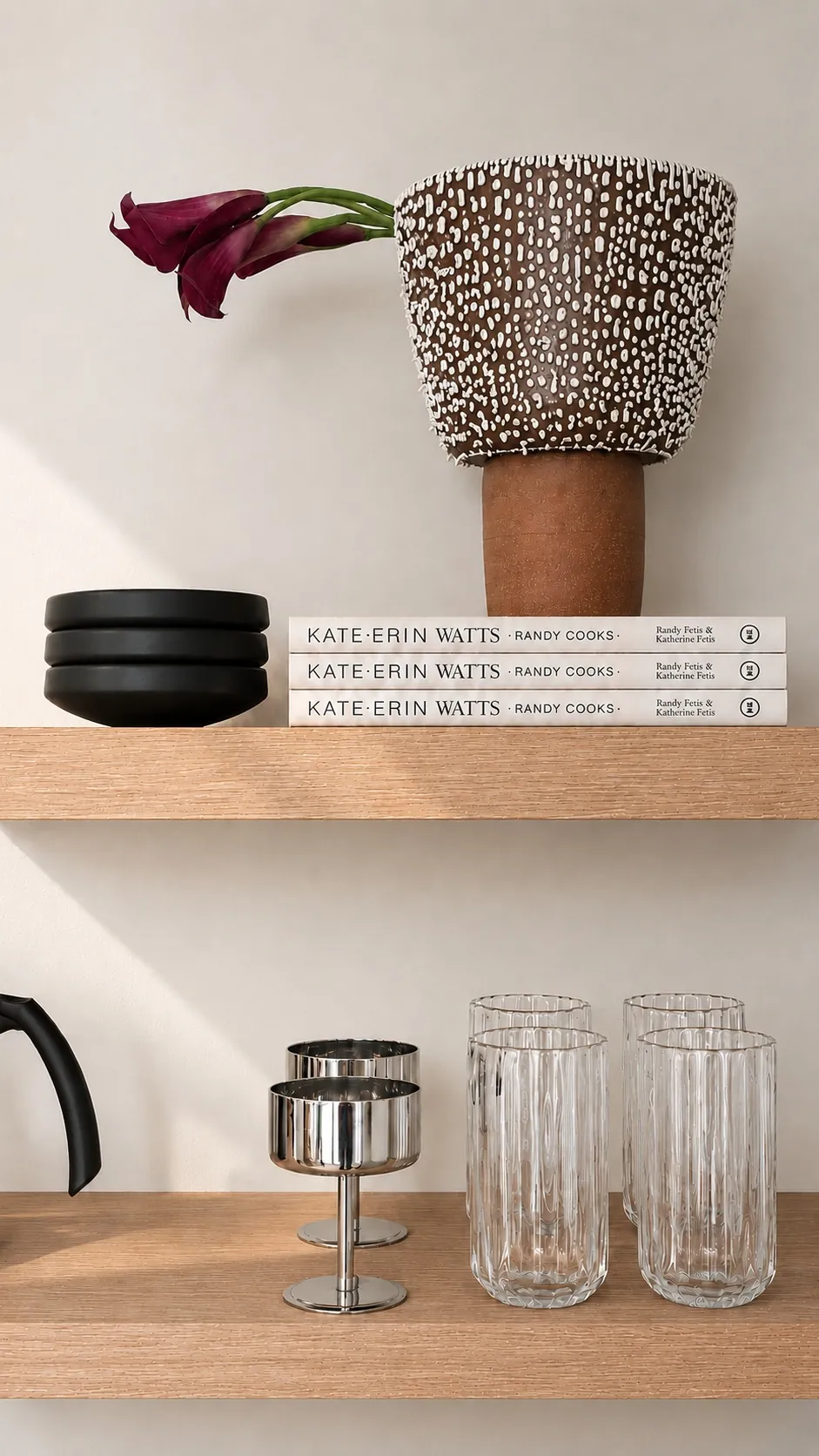 A wooden shelf displays a brown decorative vase with white dots holding a purple flower, two stacked black bowls, three stacked books, two metal goblets, four ribbed glass cups, and part of a black kettle.