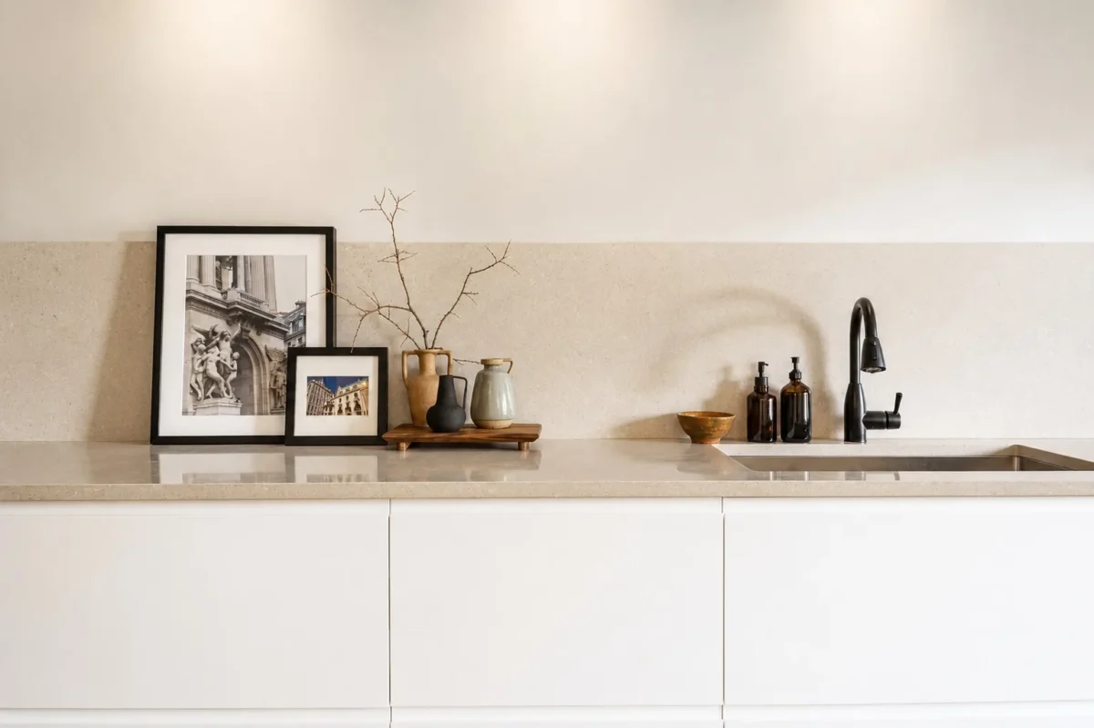 A modern kitchen counter with a black faucet, soap dispensers, a small wooden tray with vases and branches, a bowl, and two framed art photos leaning against a beige backsplash.