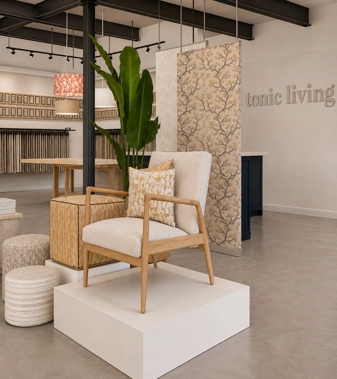 Modern showroom with a neutral armchair on a white platform, patterned pillow, round poufs, a large plant, wallpaper samples, and “tonic living” text on the wall in the background.