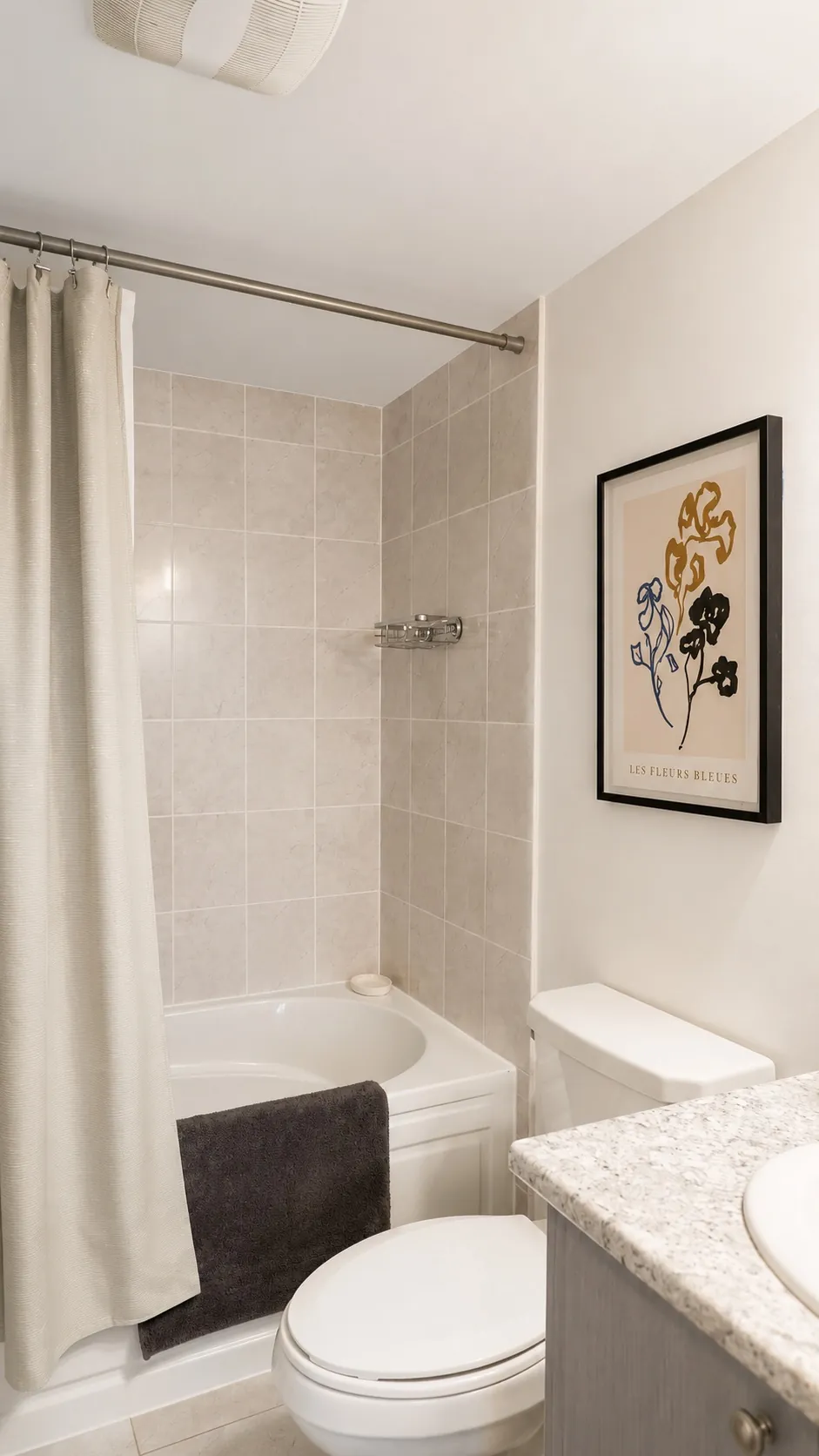 A clean, modern bathroom with beige tiled walls, a bathtub with a white shower curtain, a toilet, a granite countertop, and framed artwork reading Les Fleurs Bleues on the wall.