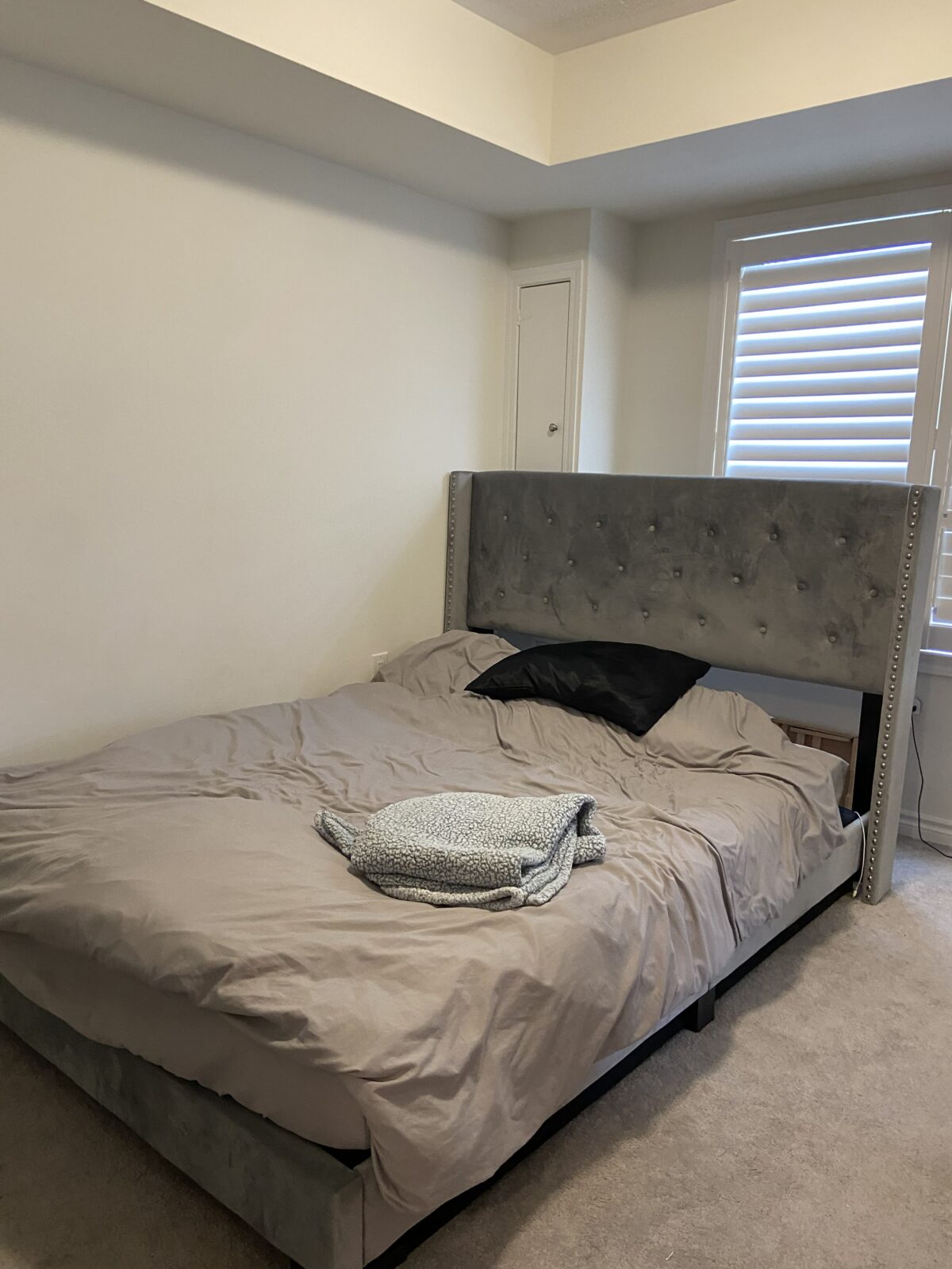 A bed with a gray upholstered headboard stands against a white wall. The bed has gray bedding, one black pillow, and a folded patterned blanket placed near the foot of the bed. A window with blinds is nearby.