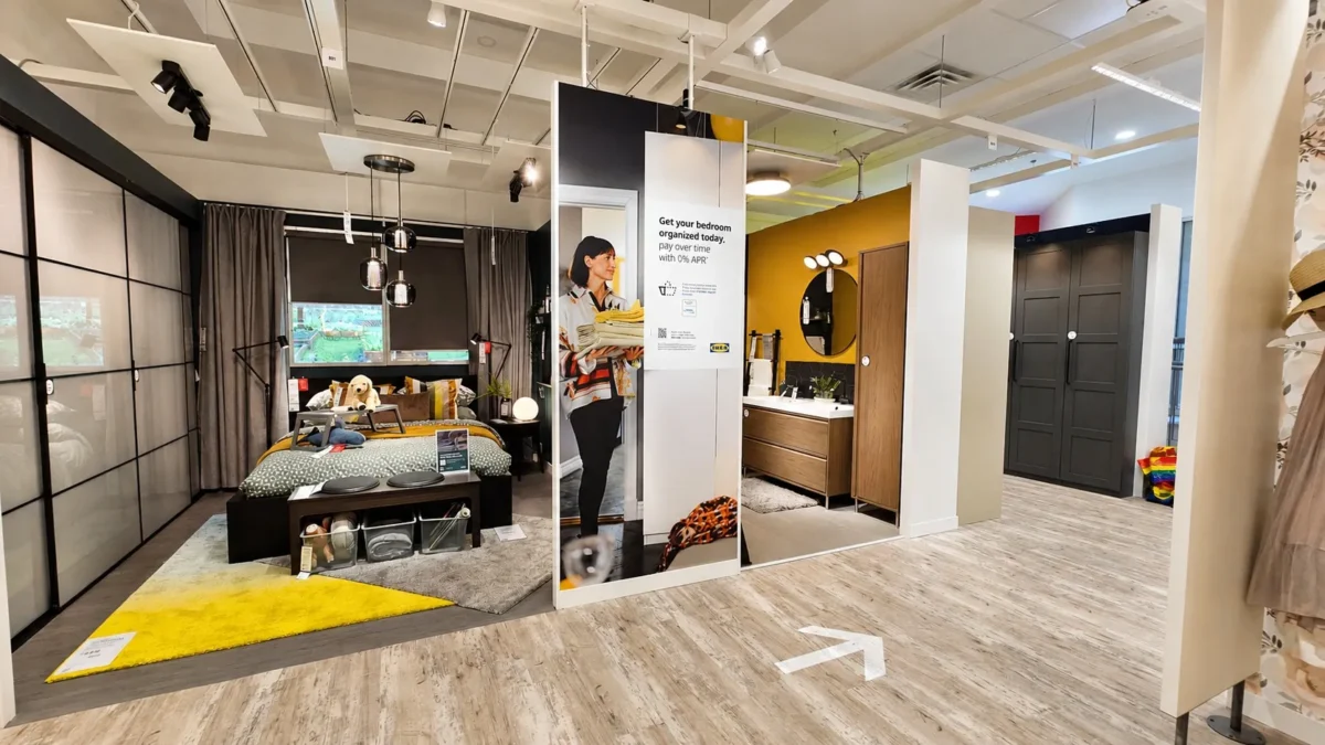 A showroom displays a modern bedroom on the left with a bed, yellow rug, and large windows, and a bathroom on the right with a round mirror and wooden cabinets. An arrow on the floor directs foot traffic.