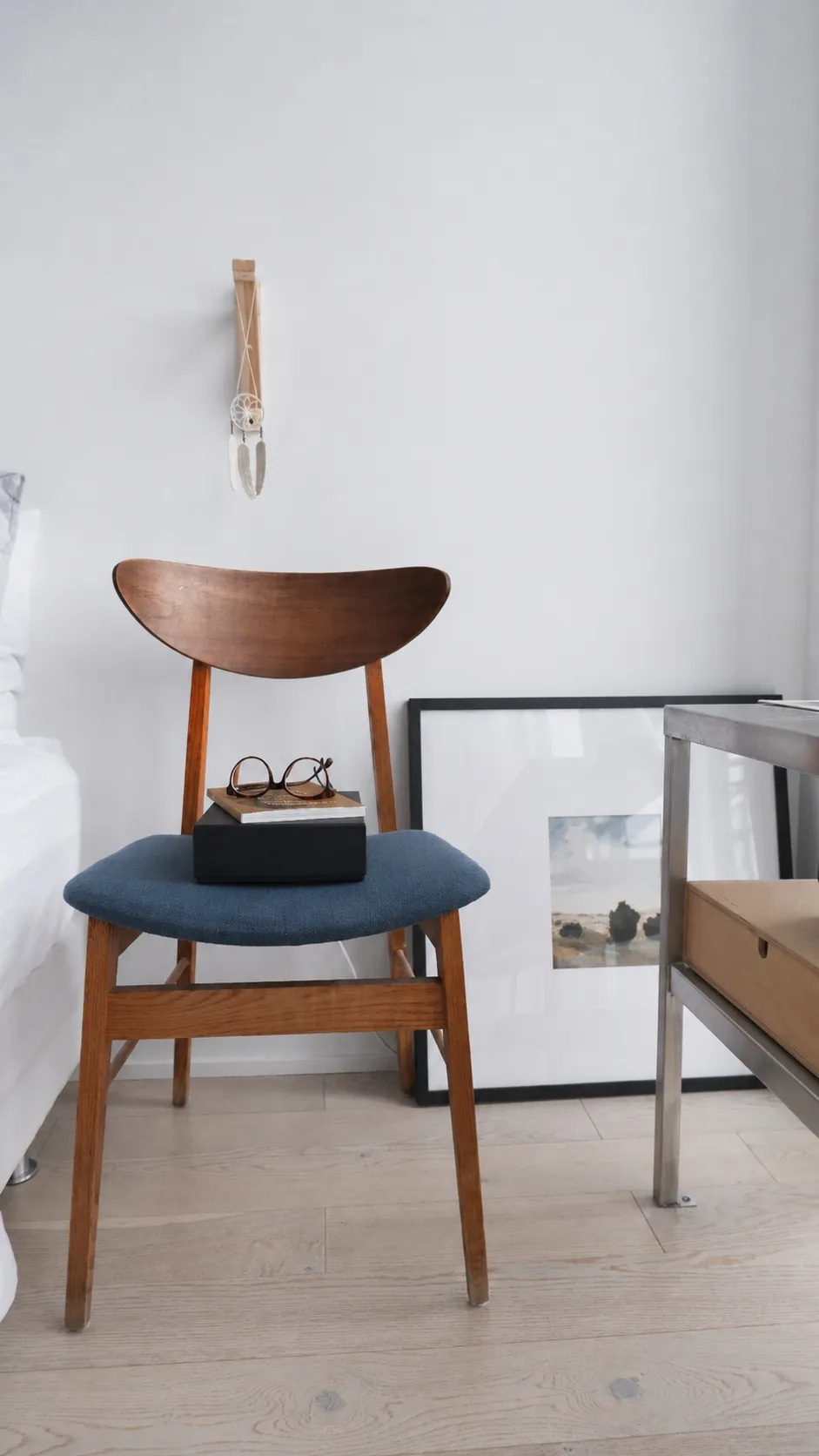 A wooden chair with a blue seat cushion holds a black book and eyeglasses. Behind it, a framed photo rests against a white wall. Part of a bed and a metal table are visible in a minimalist room.