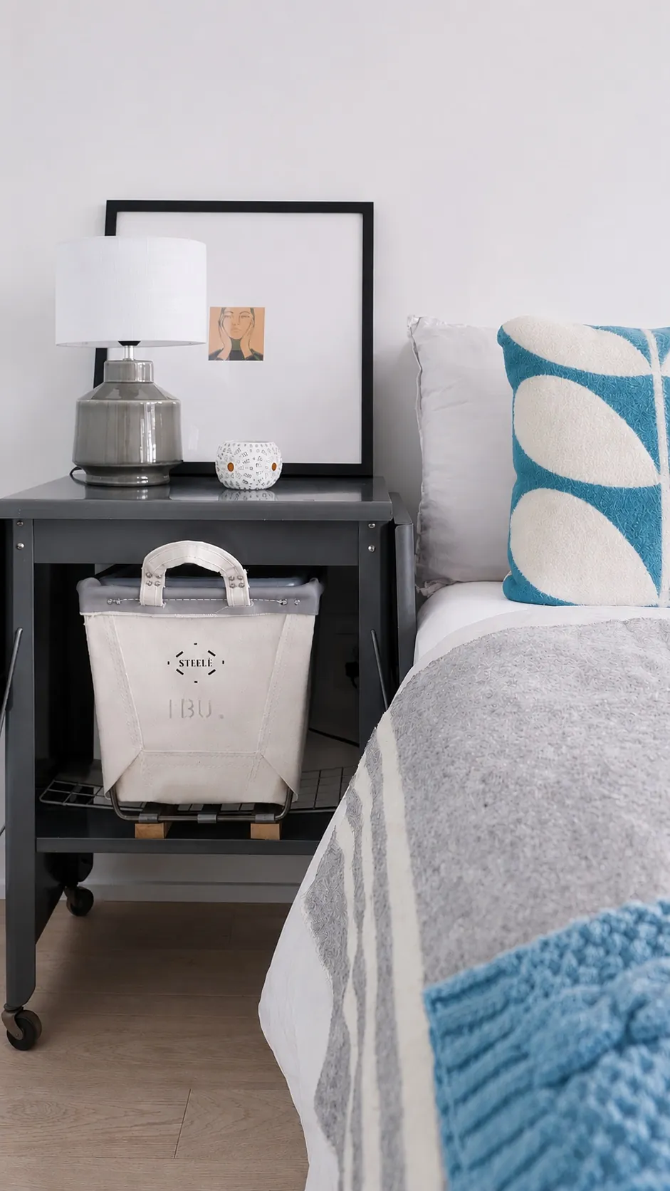 A modern bedside table holds a lamp, a framed picture, and a decorative cup. Below, a white storage bag sits on a shelf. The adjacent bed has a white and blue pillow and a gray and white blanket with a blue textured corner.