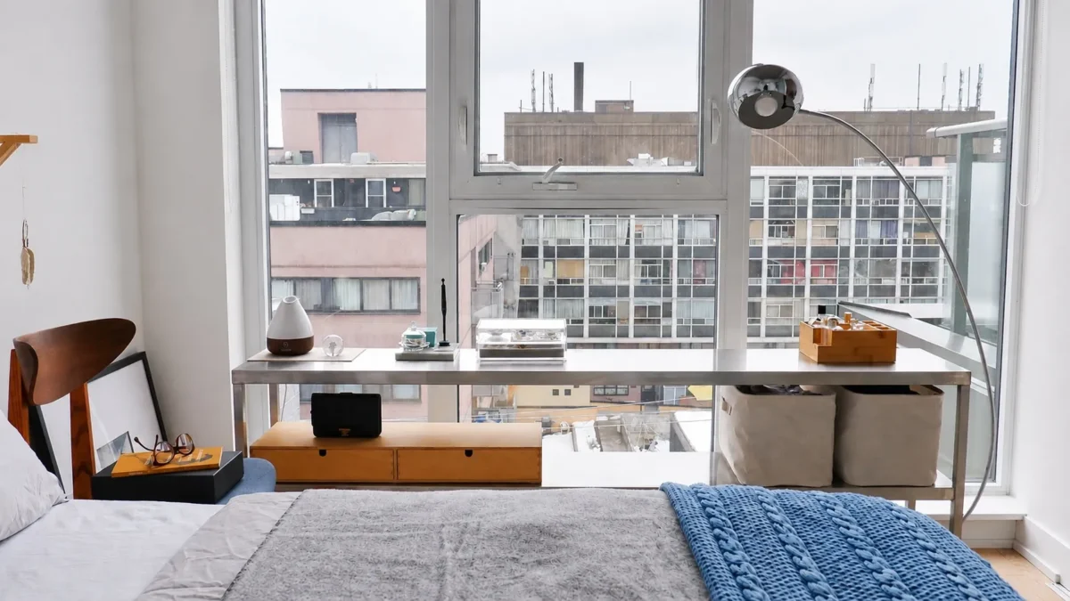 A bright bedroom with large windows, a neatly made bed, a blue knitted throw, a desk with a lamp, and organized storage bins, overlooking a cityscape of apartment buildings.