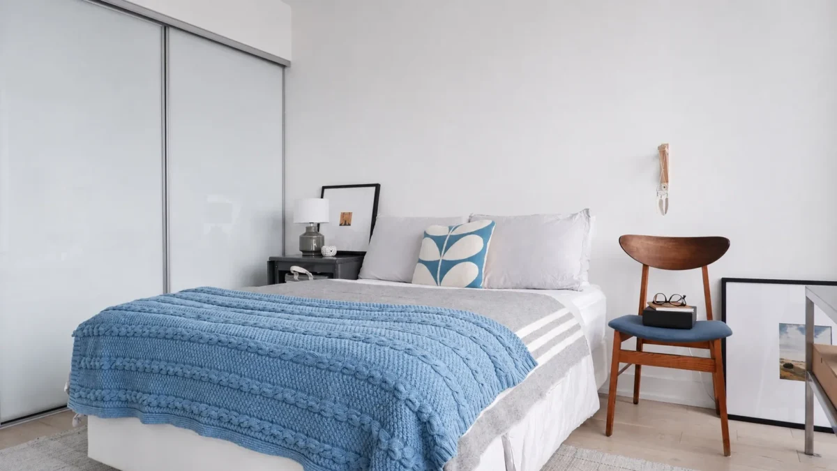 A modern, minimal bedroom features a neatly made bed with white and gray bedding, a blue textured blanket, and a leaf-patterned pillow. A nightstand, lamp, wooden chair, and framed artwork complete the decor.