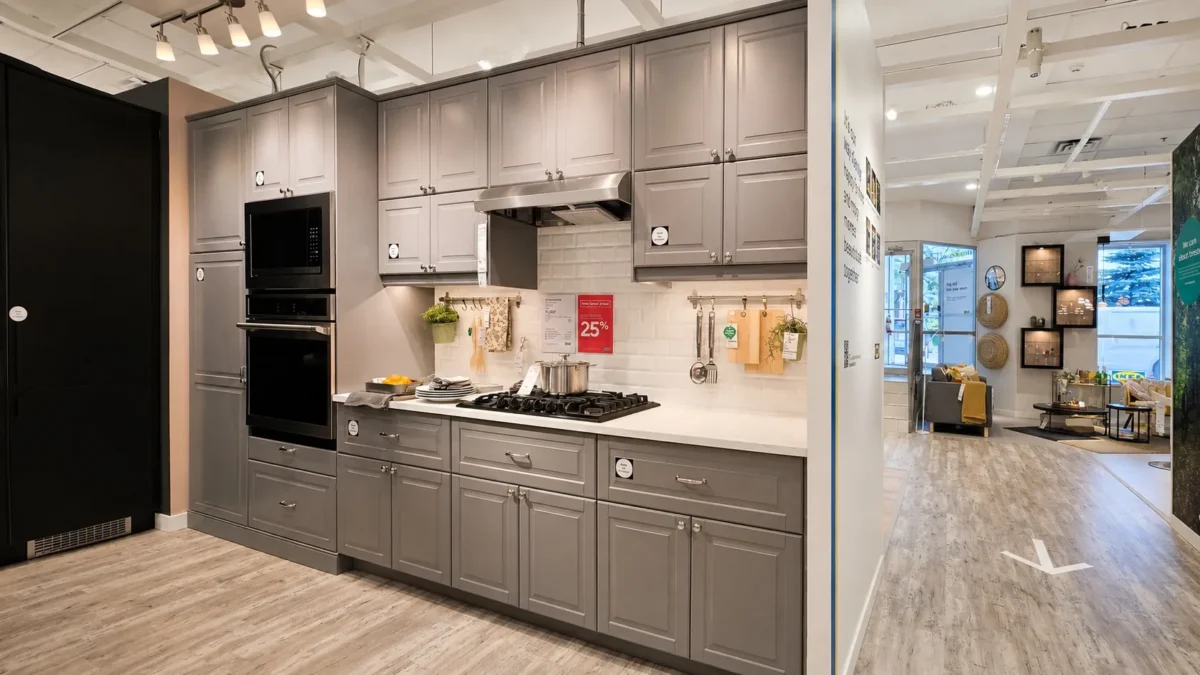 Modern kitchen with gray cabinets, white countertops, a gas cooktop, built-in oven, and under-cabinet shelves. The space has wood flooring, bright lighting, and an open layout connecting to a living area.