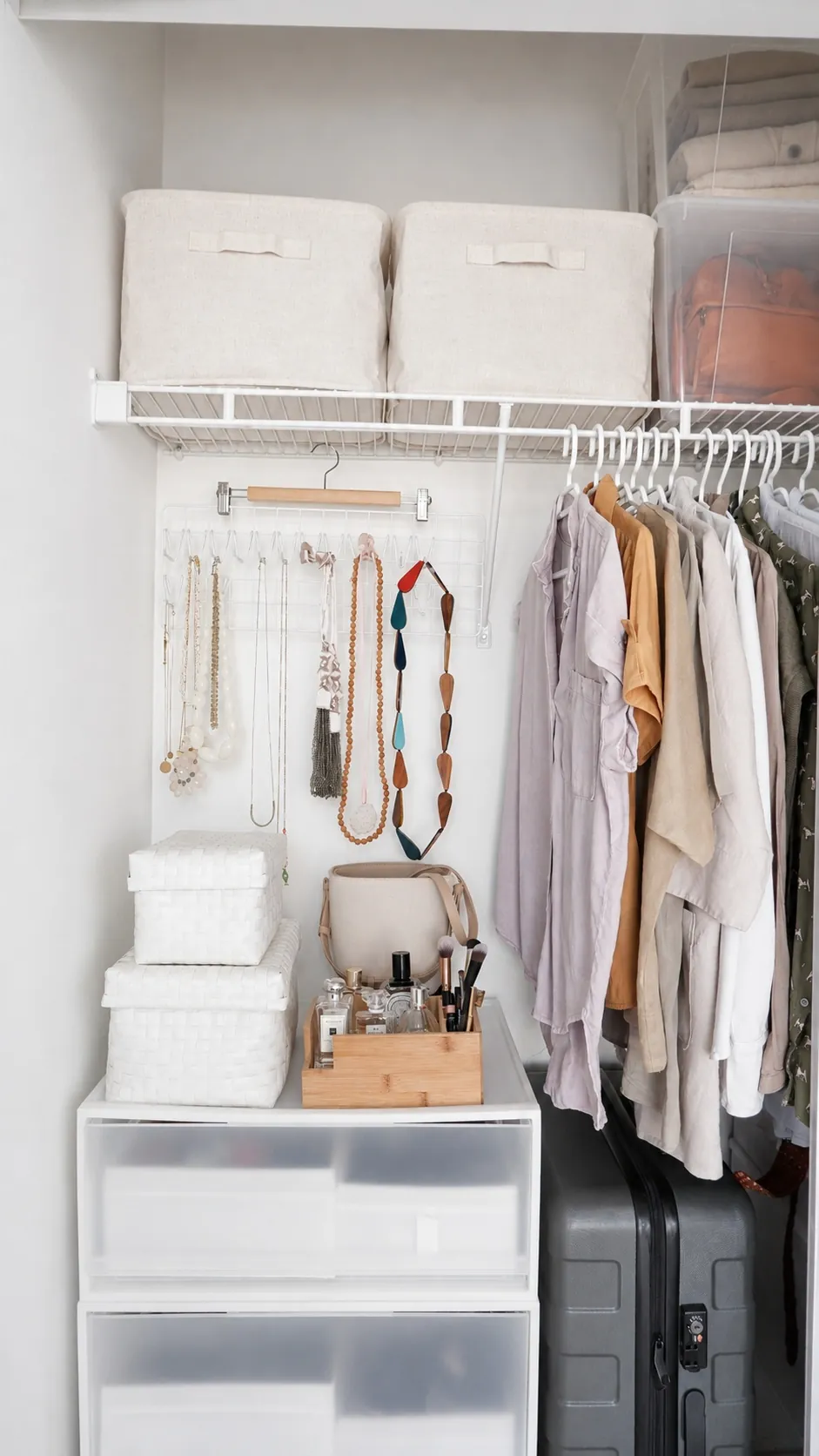 A neatly organized closet with hanging clothes, jewelry on hooks, storage bins on a wire shelf, white drawers below, a tray with beauty products, and suitcases stacked to the side.