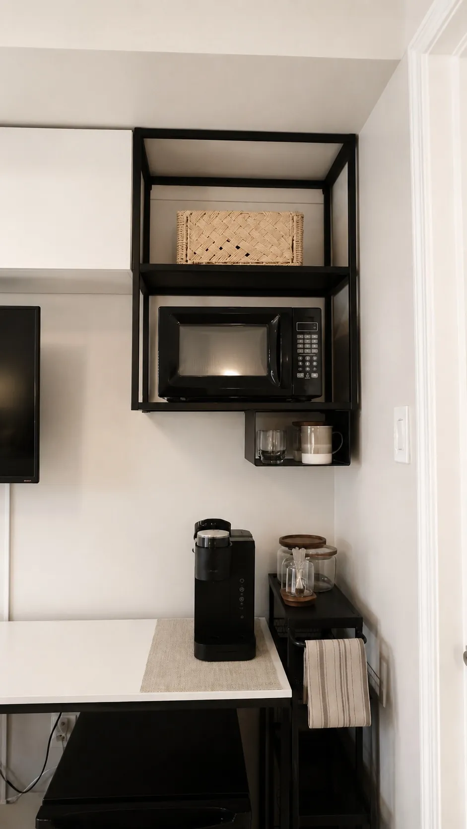 Minimalist kitchen corner with a black microwave on an open shelf, a wicker basket above, mugs and jars below, and a black coffee maker on a white counter, with a tea towel hanging on the side.
