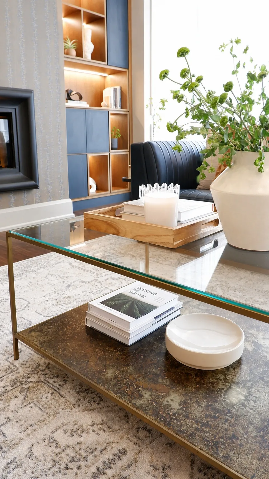 A modern living room with a glass coffee table holding books and a white bowl, a large white vase with green foliage, and a wooden tray with books on a beige rug near a navy sofa and built-in shelving.