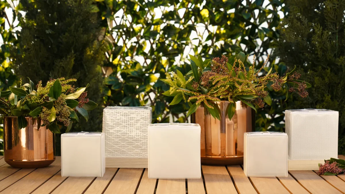 Potted plants in copper containers and four white tissue boxes of different sizes are arranged on a wooden table outdoors, with green leafy shrubs in the background.