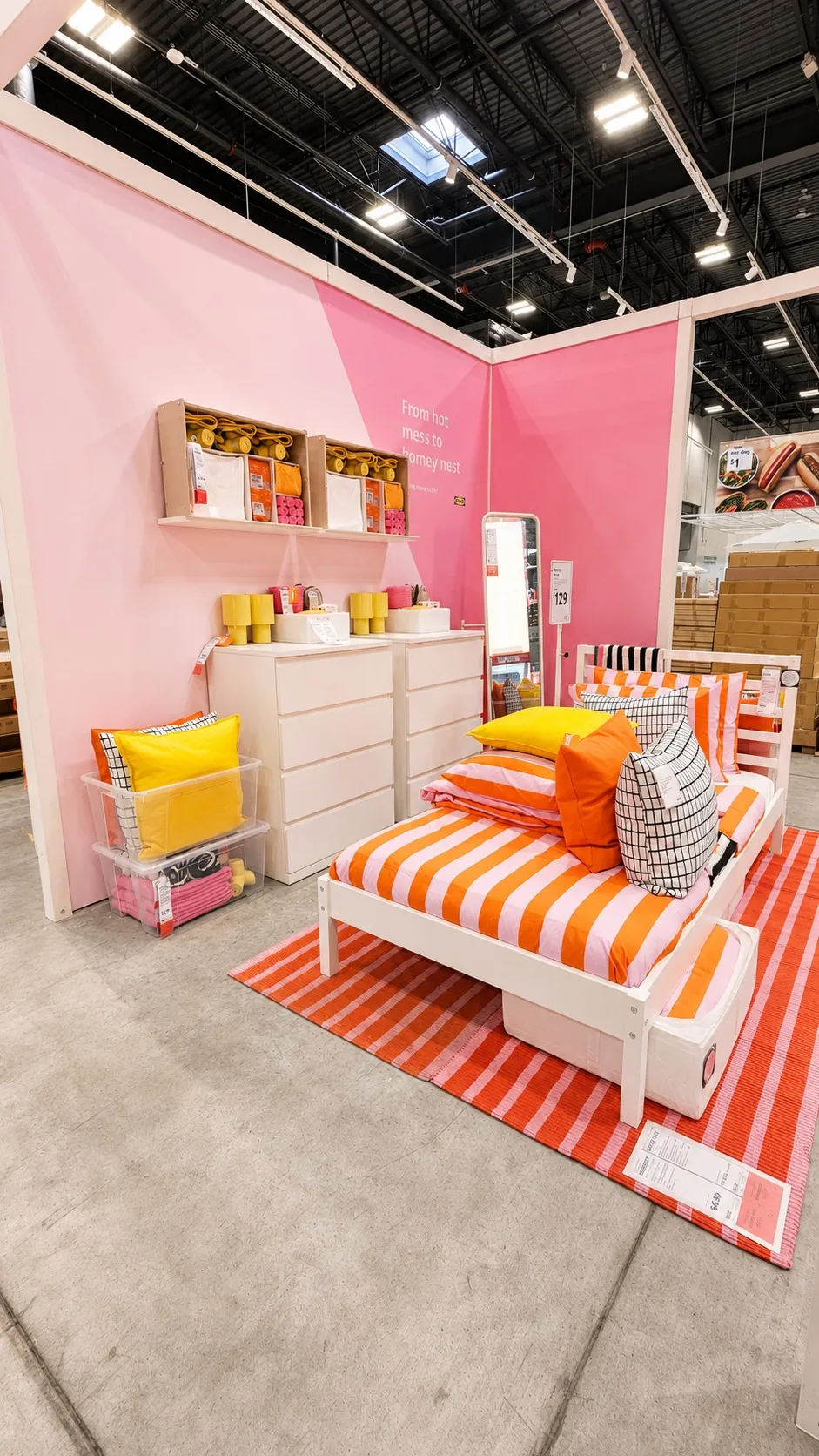 A showroom display features a bed with orange and white striped bedding and colorful pillows, a striped rug, white dressers, shelves with decor items, and a pink accent wall in a modern, well-lit store.