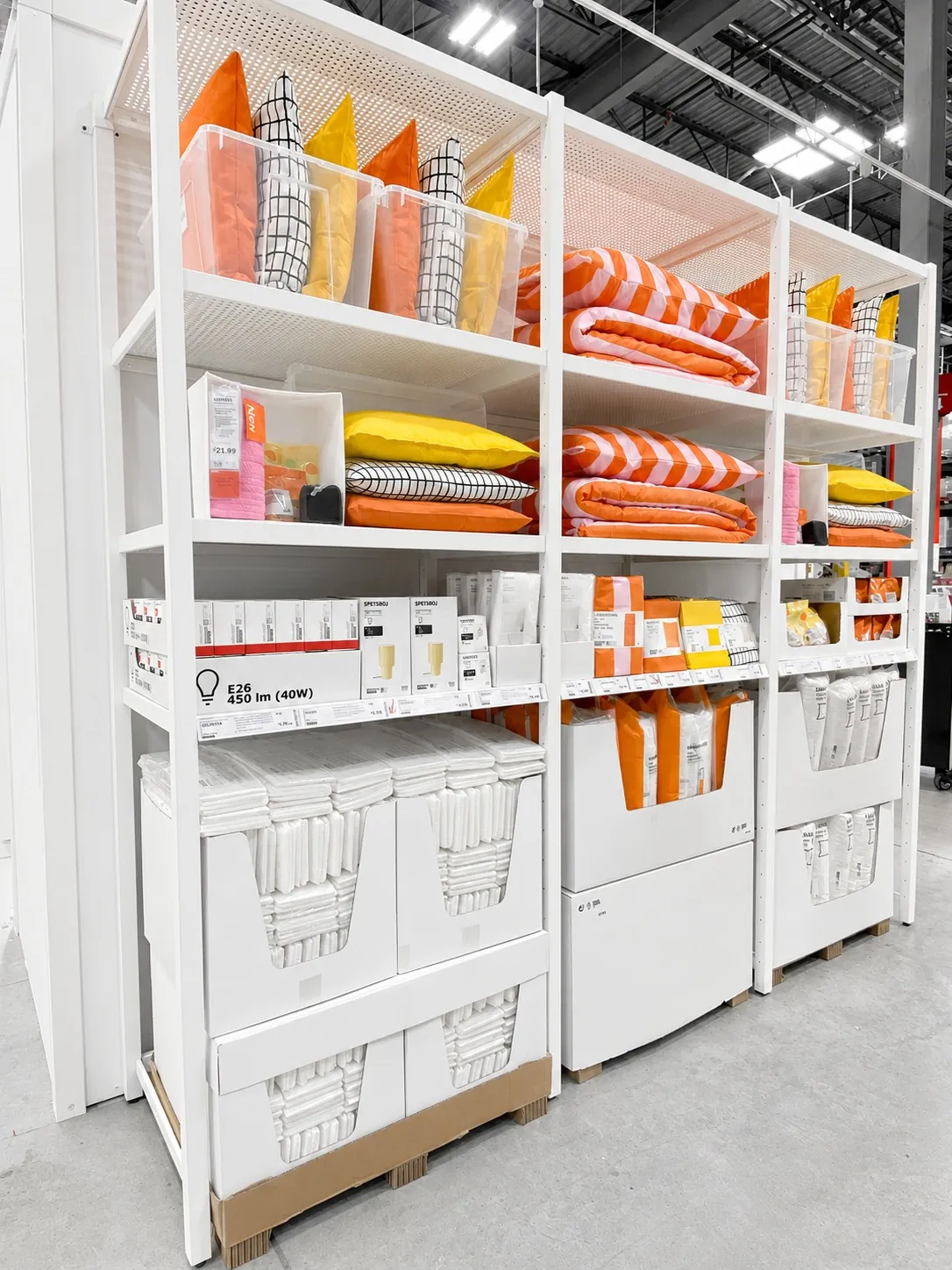 White shelving unit displays colorful pillows, throws, and home accessories in orange, yellow, and black patterns. Lower shelves hold boxed light bulbs and baskets, all arranged neatly in a brightly lit store.