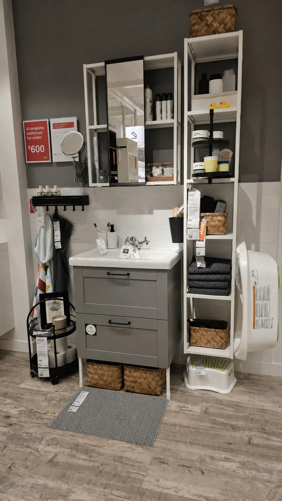 A modern bathroom display with a gray vanity, white sink, open shelving stocked with towels, baskets, and toiletries, a wall mirror, and a round laundry hamper on a wooden floor. Price tags are visible on the items.