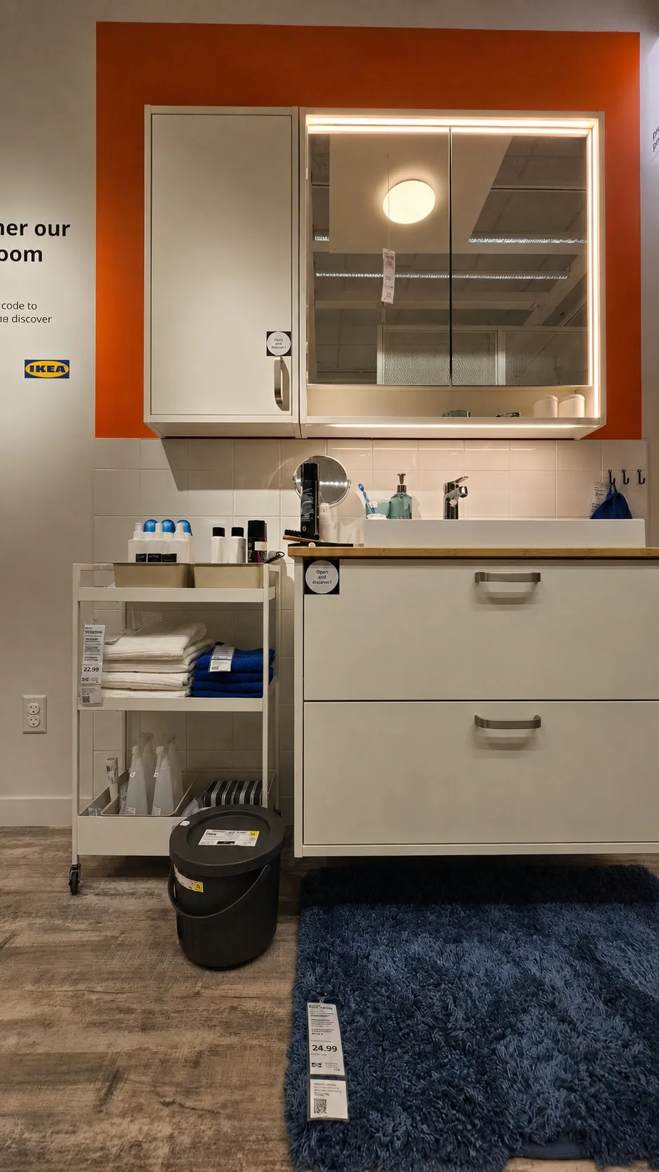 A modern bathroom display at IKEA with white cabinets, a mirrored medicine cabinet, toiletries, neatly folded towels, a blue rug, and a trash bin on a wood-patterned floor. Orange accent paint frames the cabinet.
