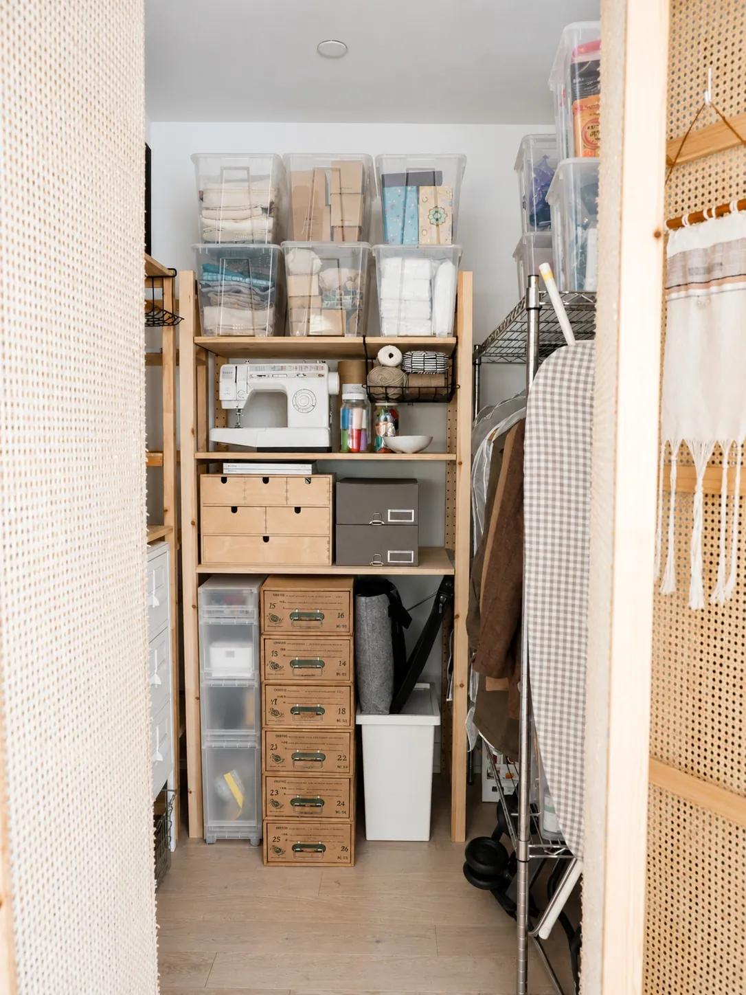 A well-organized closet with shelves holding clear storage bins, a sewing machine, drawers, baskets, craft supplies, and hanging clothes. An ironing board and laundry basket are also visible.