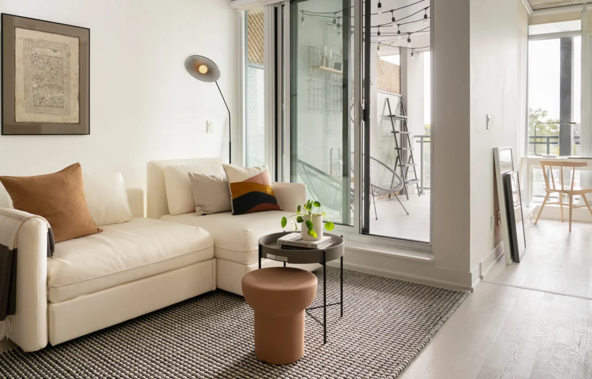 A modern living room with a white sectional sofa, brown and beige pillows, a small round table with a plant, a gray rug, and glass doors opening to a balcony with string lights and outdoor seating.