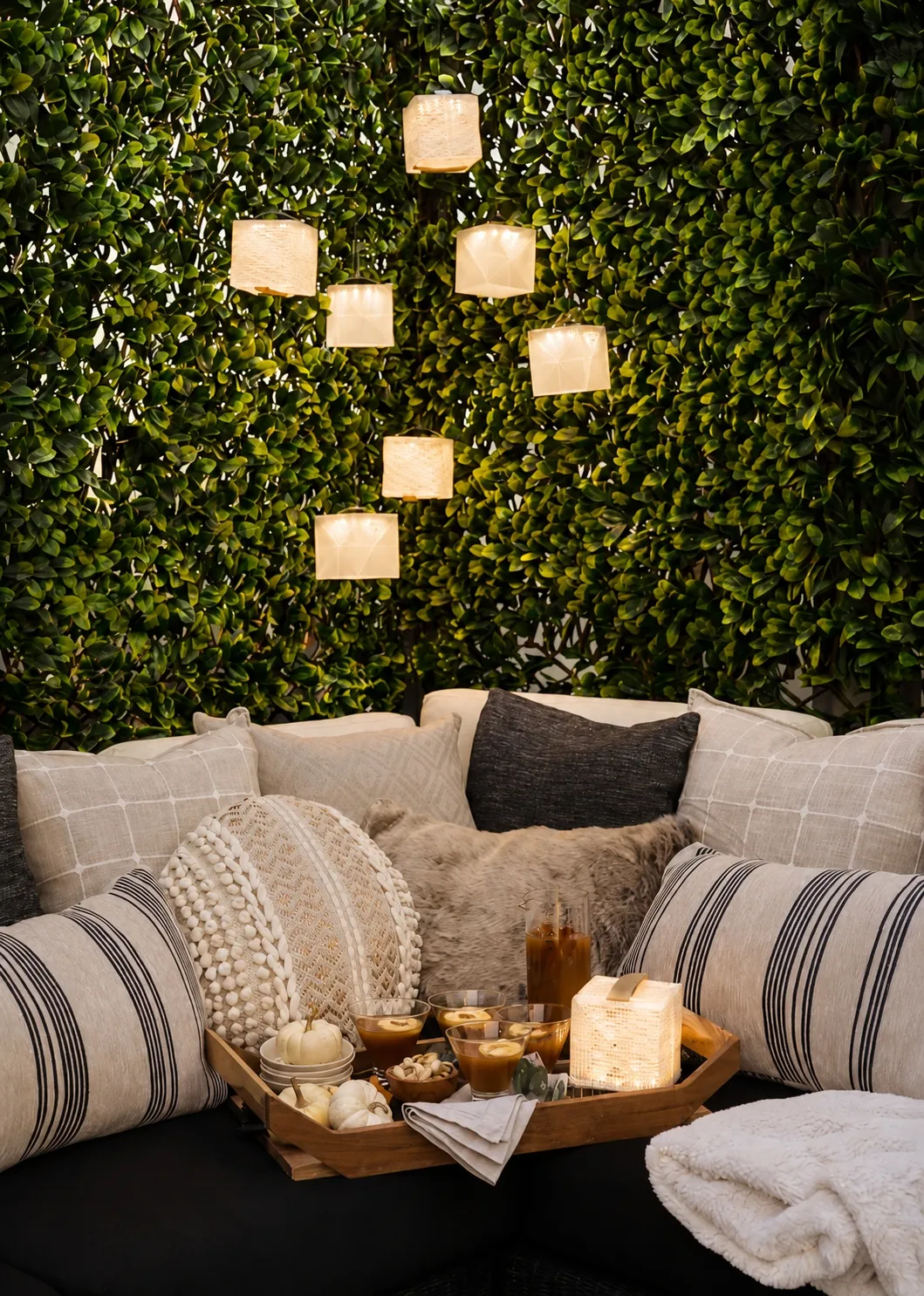 A cozy outdoor seating area with patterned and textured pillows, a furry throw blanket, a green leafy wall, and soft cube lantern lights. A wooden tray holds snacks, drinks, small pumpkins, and a glowing lantern.