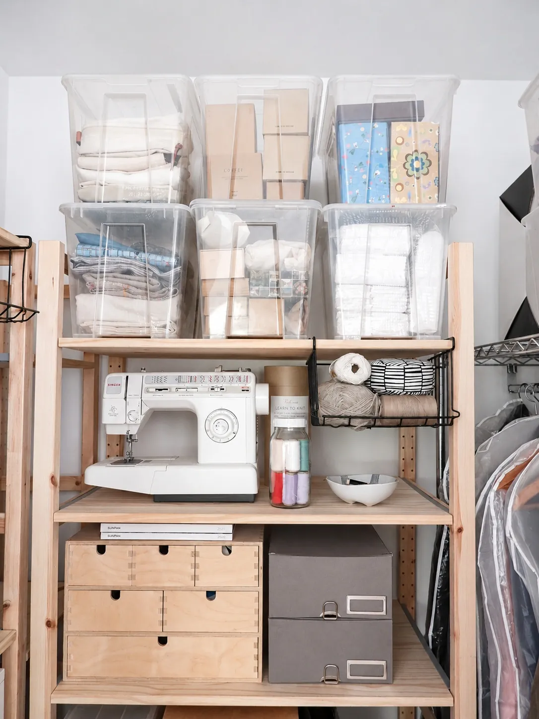 A neatly organized wooden shelving unit holds clear plastic bins with fabric and boxes, a sewing machine, thread spools, a bowl, and gray and wooden storage boxes. Garment bags hang to the right.
