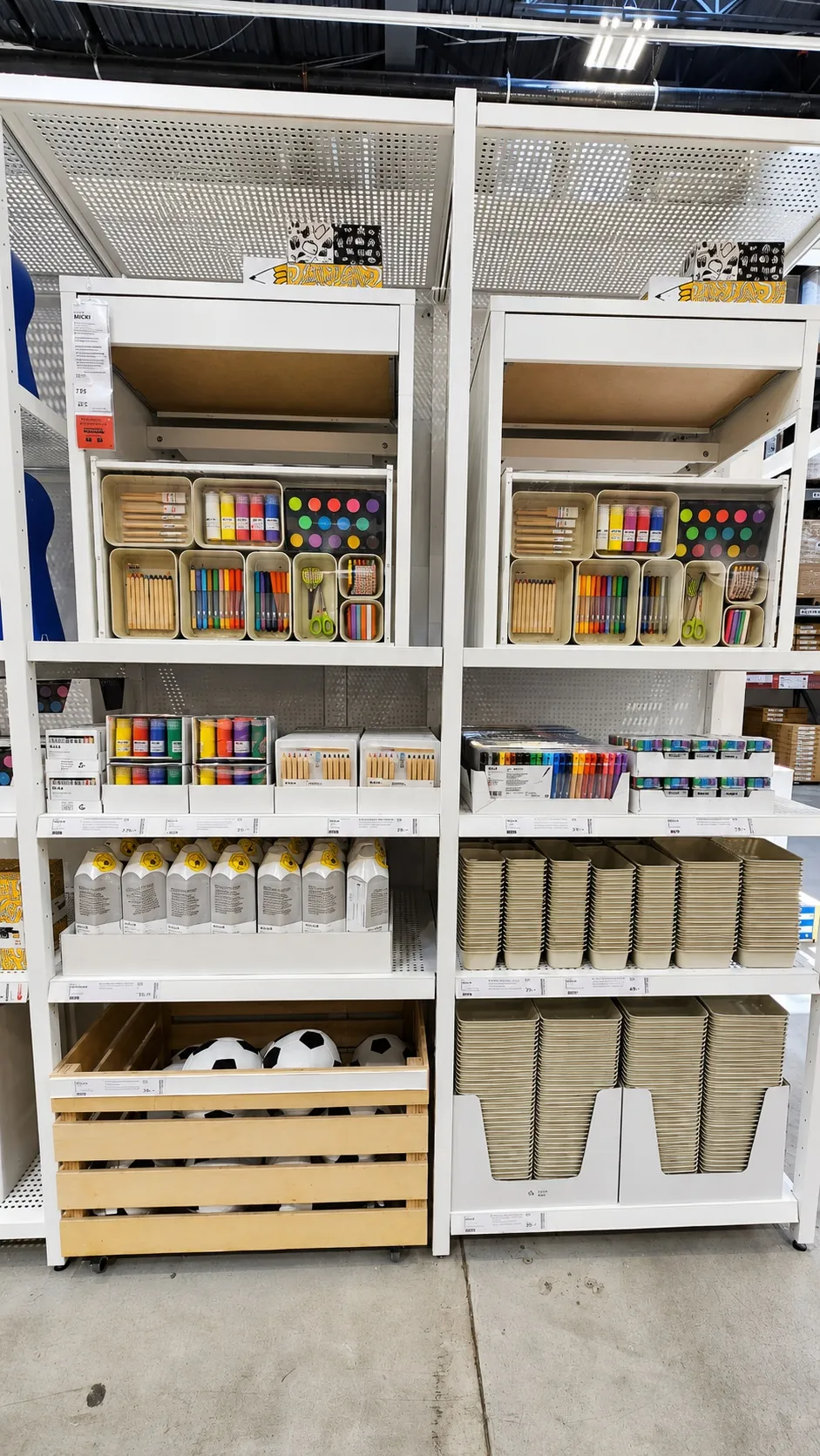 Shelves display various school and craft supplies, including colored paints, markers, brushes, colored pencils, sketchpads, and white bottles. A wooden crate at the bottom holds several white soccer balls.