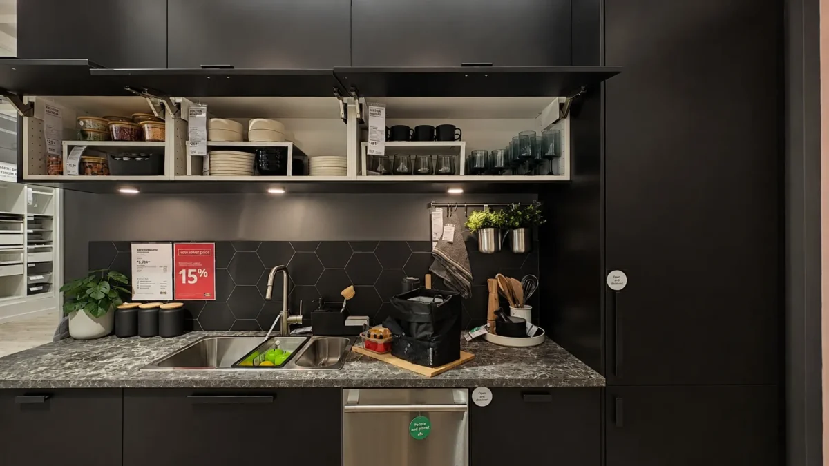A modern black kitchen with open upper cabinets displaying dishes, glasses, and mugs. Below, a marble countertop holds a sink, cutting board, kitchen tools, and small plants. A dishwasher and under-cabinet lighting are visible.