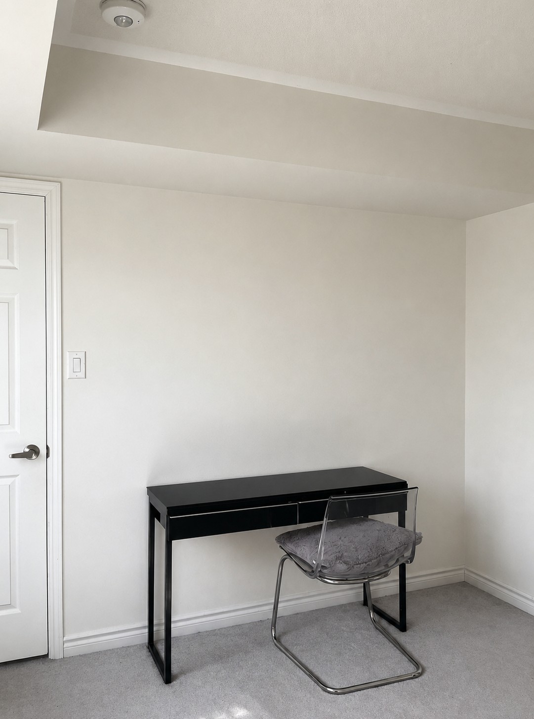 A minimalist room with a small black desk and a transparent chair with a gray cushion, placed against a white wall near a closed white door. The carpet is light gray, and the space is well-lit.
