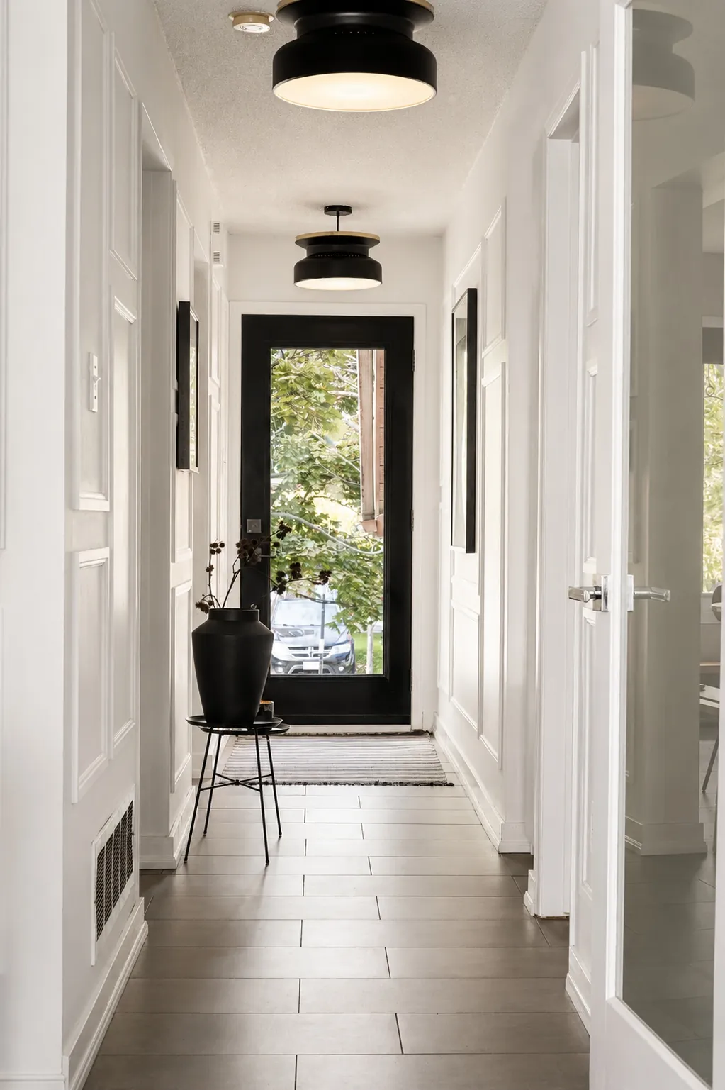 A bright hallway with white walls and light tile flooring leads to a glass door. A black planter with a plant sits on a stand near the door, and two modern black ceiling lights hang above. Natural light fills the space.