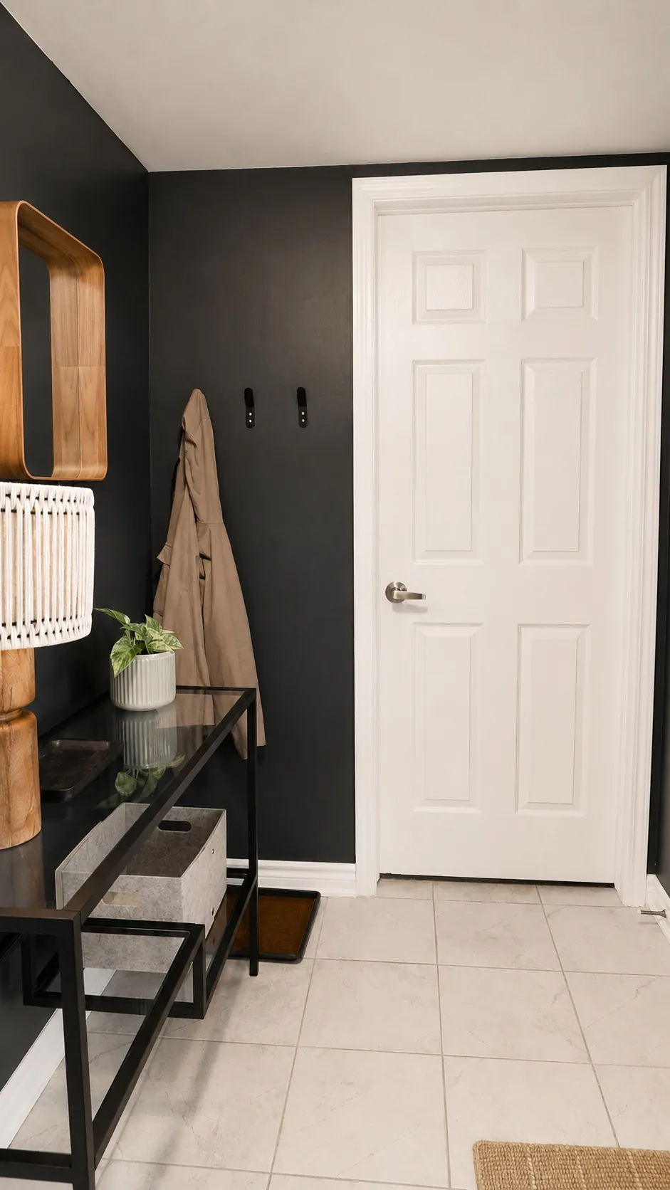 Modern entryway with a white door, black walls, a tan coat hanging on wall hooks, a black console table with a potted plant, basket, and boxes, and light tile flooring.