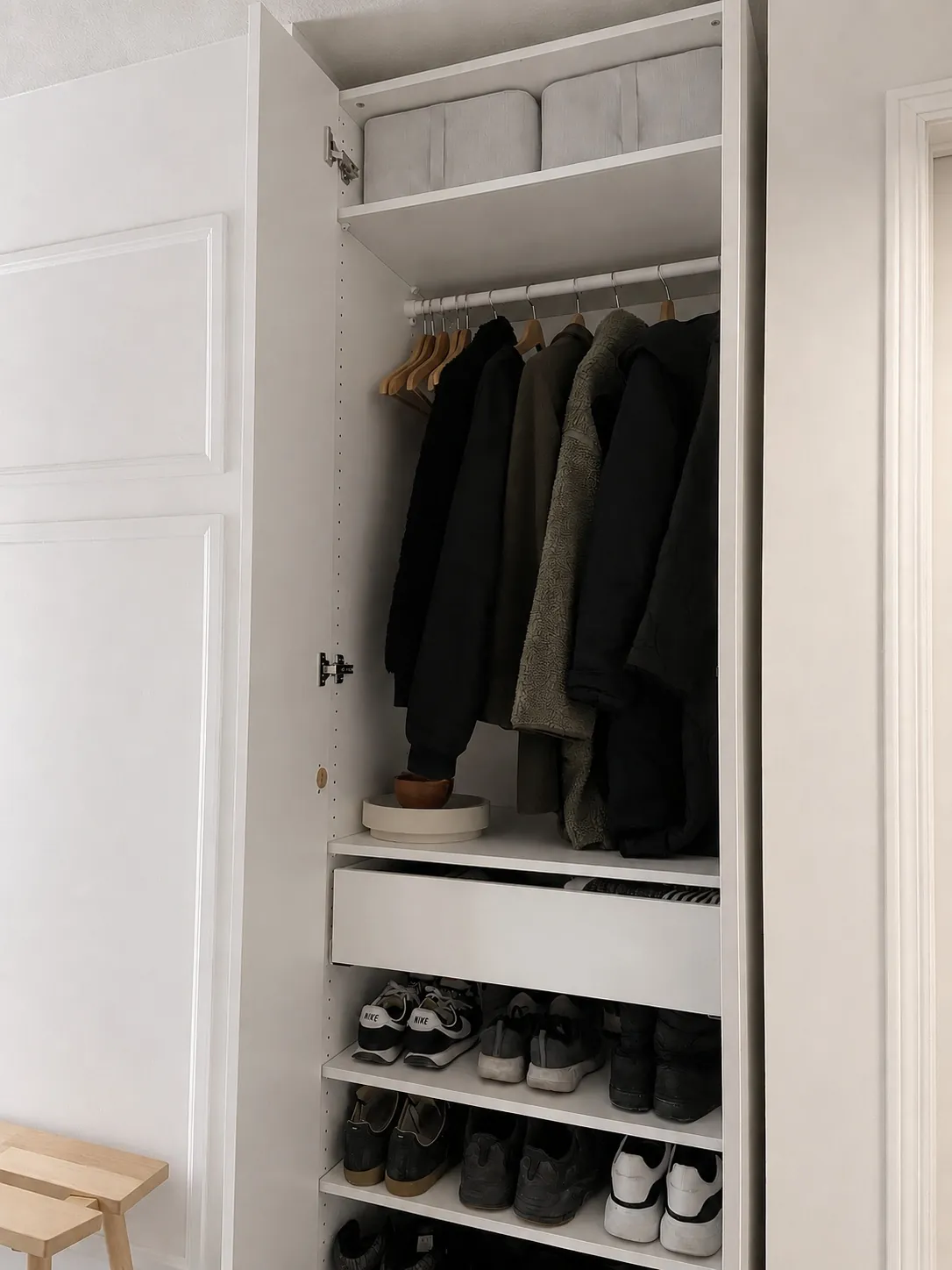 Open white wardrobe with neatly hung jackets on wooden hangers, two gray storage boxes on the top shelf, a hat and tray on a middle shelf, and shoes organized on three lower shelves.
