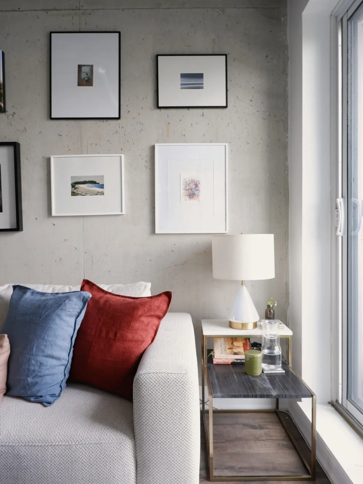 A modern living room with a light gray sofa, blue and red pillows, a small side table holding a lamp, a glass of water, and a green mug, with framed art on a concrete wall and a large window letting in natural light.