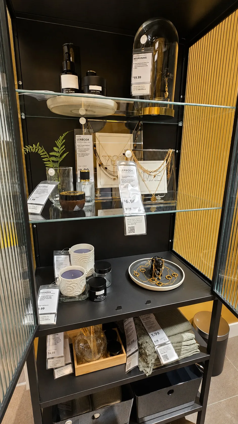 A black display cabinet with glass shelves holding candles, jewelry, framed artwork, decorative accessories, folded textiles, and price tags in a modern store setting with a yellow-striped wall background.