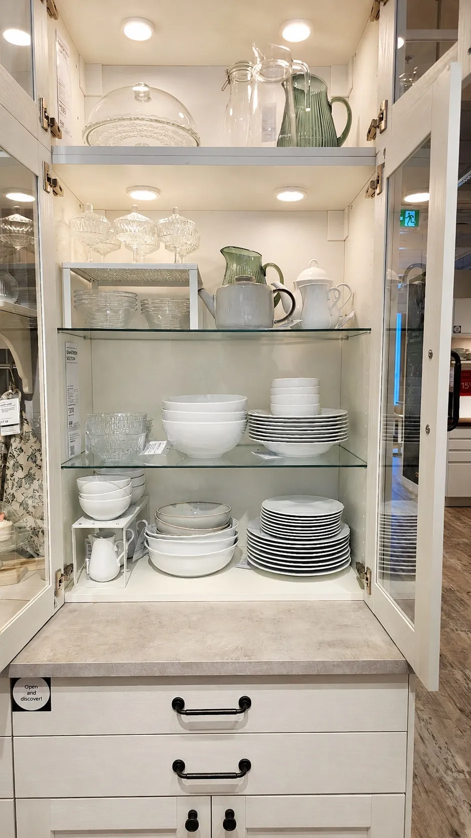 A glass-fronted kitchen cabinet displaying neatly arranged white dishes, bowls, mugs, glassware, and pitchers on glass shelves, with overhead lighting illuminating the items.