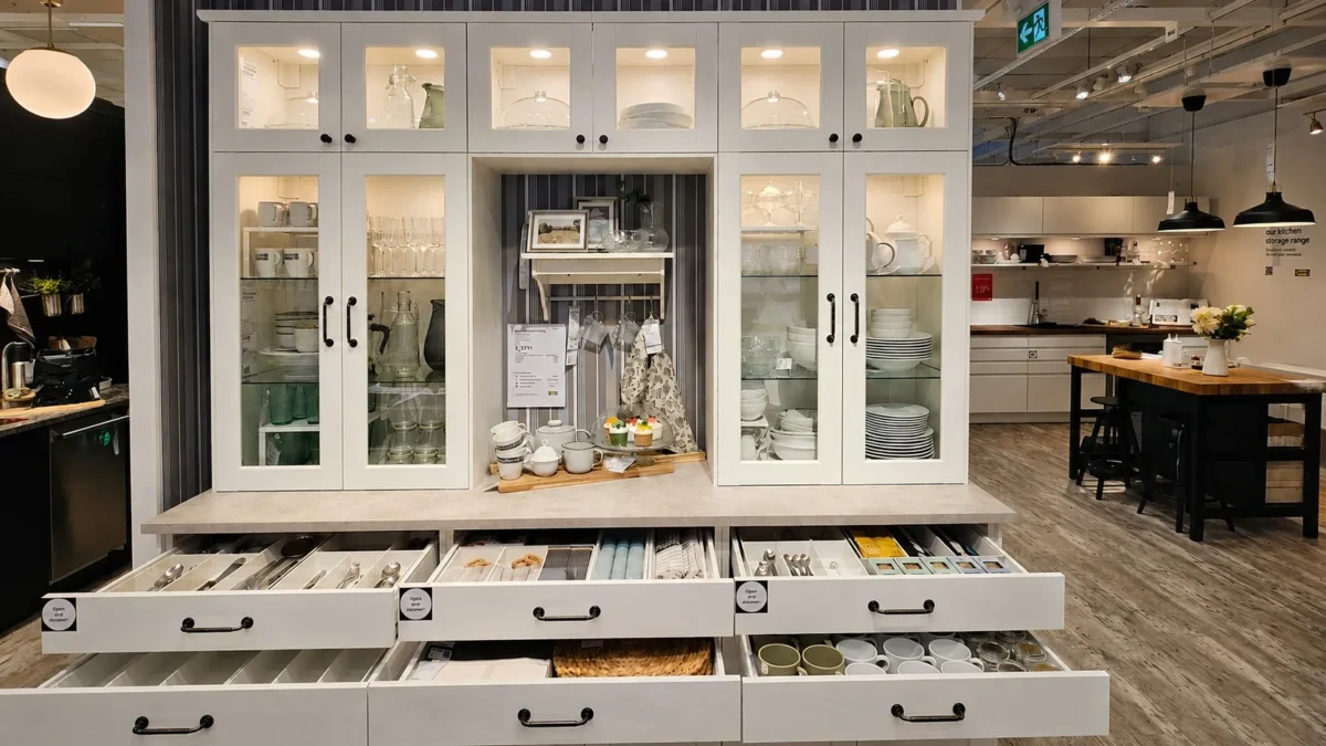 A white kitchen hutch with glass-front cabinets displays dishes and glassware. The countertop holds kitchen decor, while open drawers below reveal cutlery, utensils, and linens. The scene is in a modern showroom.