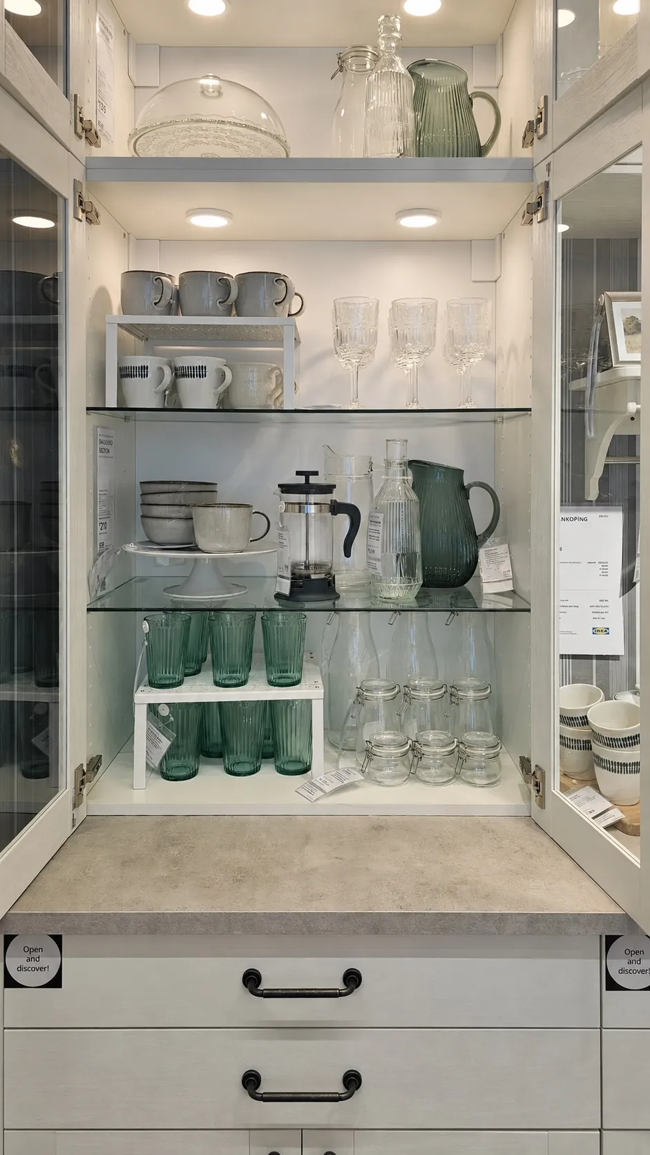 A glass-front cabinet displays neatly arranged mugs, bowls, glassware, a French press, a pitcher, and green drinking glasses on white shelves with interior lighting.