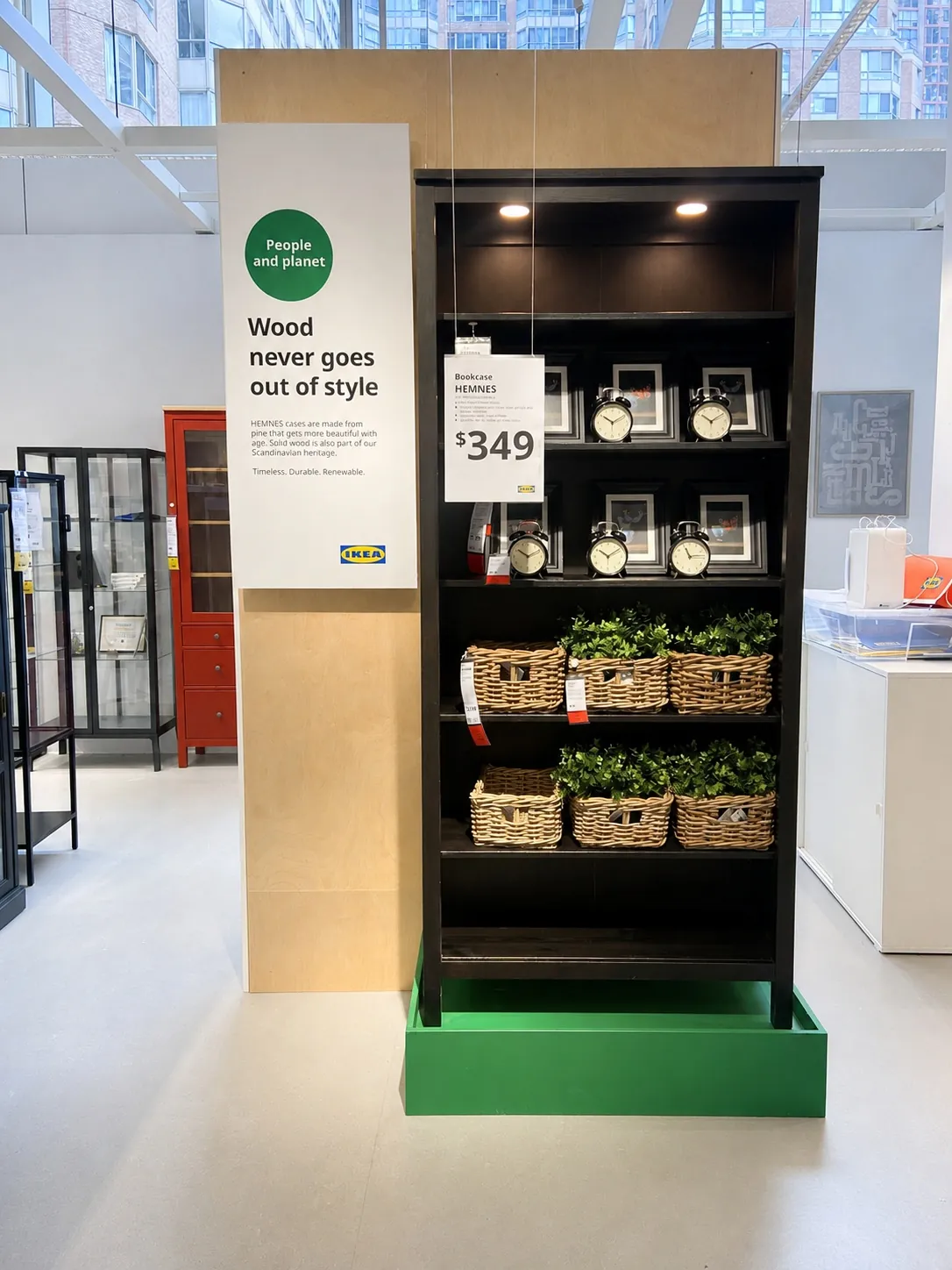 A black shelving unit on display at a store holds clocks and wicker baskets with green plants. A sign beside it reads, Wood never goes out of style.
