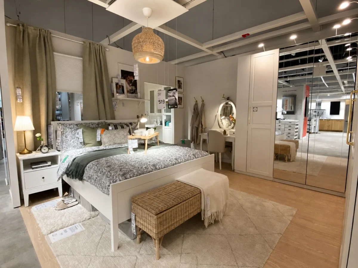 A cozy, modern bedroom display in a furniture store features a neatly made bed with patterned bedding, nightstands with lamps, a wicker bench, mirrored wardrobe, vanity, and warm lighting.